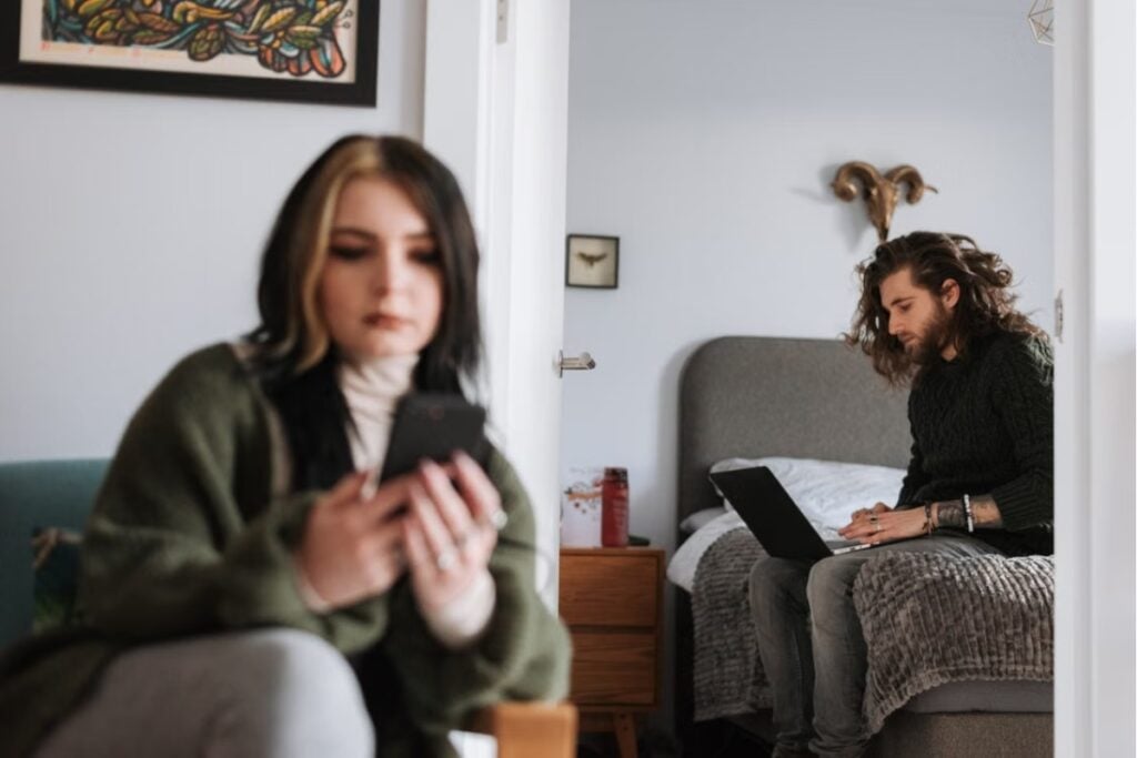 Fotografia de plano médio mostrando uma mulher em primeiro plano desfocado sentada olhando para o celular, enquanto ao fundo, através da porta aberta do quarto, um homem com cabelos compridos trabalha no notebook sentado na cama.