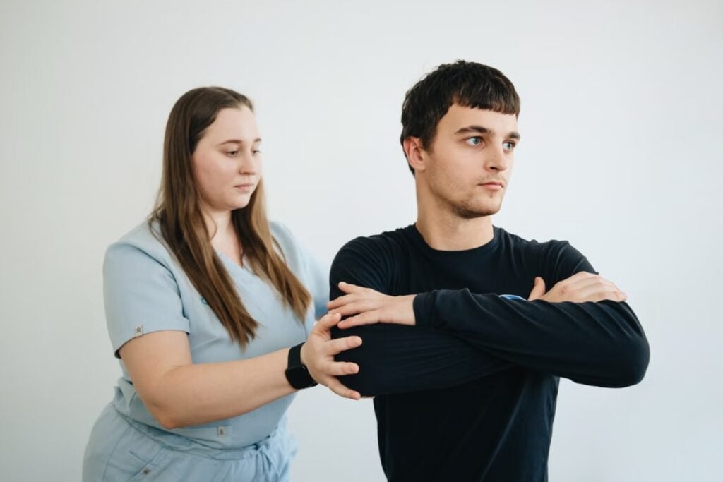 Fotografia de plano médio mostrando um homem de camiseta preta com os braços cruzados sobre o peito, enquanto uma profissional de saúde de uniforme azul-claro segura seu cotovelo para realizar um teste ou ajuste postural diante de um fundo branco.