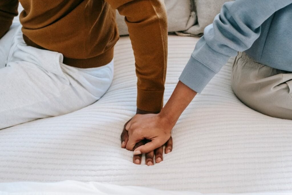 Fotografia em plano detalhe aproximado mostrando as mãos entrelaçadas de um casal, uma de pele escura e outra de pele mais clara, apoiadas sobre um colchão ou colcha branca texturizada, enquanto ambos estão sentados próximos vestindo blusas de moletom em tons de marrom e azul-claro.