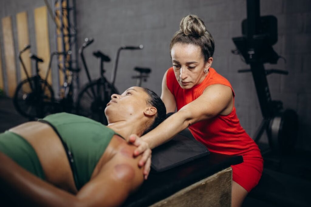Fotografia de plano médio de uma terapeuta de vestido vermelho e cabelo preso, inclinada sobre uma atleta deitada de costas que veste top verde e apresenta marcas redondas de ventosaterapia no ombro, realizando uma manobra de descompressão no pescoço em um ambiente de academia ao fundo.