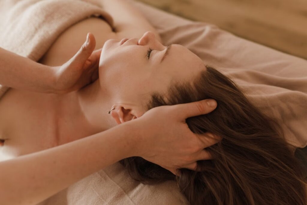 Fotografia em plano detalhe e ângulo superior de uma mulher de olhos fechados deitada de costas em uma maca, recebendo uma massagem suave na mandíbula e no couro cabeludo pelas mãos de uma terapeuta, em um ambiente com tons pastéis e acolhedores.