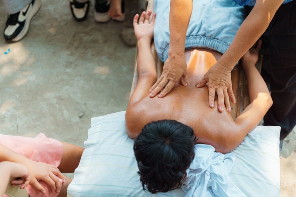 Fotografia em ângulo superior (plano zenital parcial) mostrando um jovem deitado de bruços em um banco de madeira improvisado com um travesseiro, recebendo uma massagem com óleo nas costas aplicada por duas mãos firmes, enquanto outras pessoas observam ao redor em um chão de terra batida.