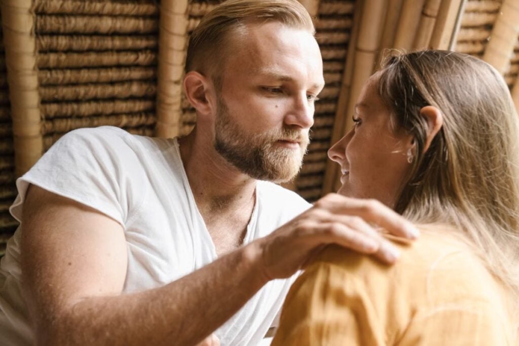 Fotografia em plano médio aproximado mostrando um homem loiro com barba, vestindo camiseta branca, olhando fixamente nos olhos de uma mulher de perfil enquanto repousa a mão carinhosamente sobre o ombro dela, em ambiente com teto de palha ou bambu.