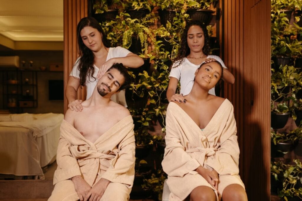 Fotografia de plano médio mostrando um homem e uma mulher sentados lado a lado de roupão bege, com os olhos fechados e a cabeça inclinada para o lado, enquanto duas terapeutas de camiseta branca realizam massagens em seus pescoços e ombros diante de um painel de plantas.