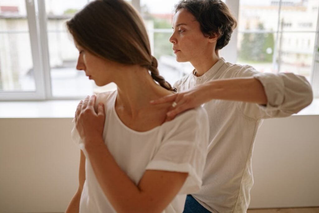 Fotografia de plano médio mostrando uma mulher de camiseta branca em primeiro plano de perfil, enquanto outra mulher ao fundo, vestindo camisa clara, posiciona as mãos em seus ombros para iniciar uma manobra de massagem ou ajuste postural em um ambiente iluminado por janelas.