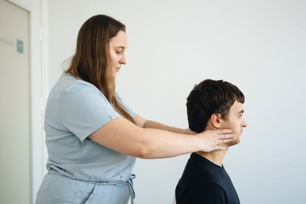 Fotografia de plano médio mostrando o perfil de um homem jovem sentado de camiseta preta, enquanto uma terapeuta em pé, vestindo uniforme médico azul-claro, apoia as mãos na lateral e na base do pescoço dele para realizar um exame ou procedimento clínico, contra uma parede branca.
