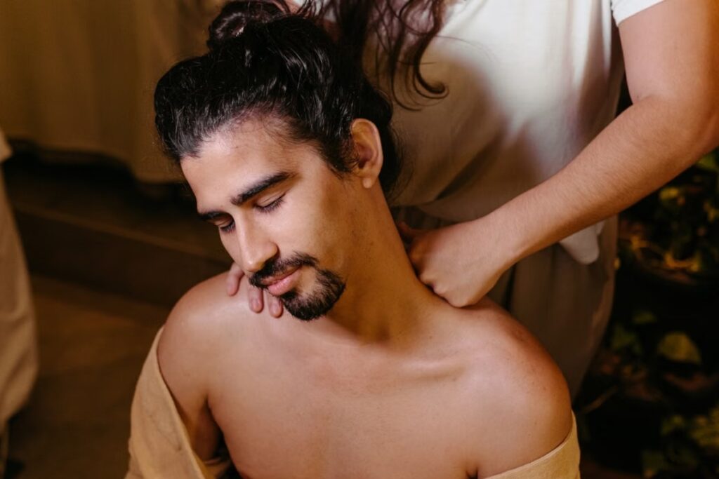 Fotografia em plano médio de um homem de olhos fechados e expressão serena com o cabelo preso, recebendo uma massagem no pescoço e ombros pelas mãos de uma terapeuta vestida de branco, em um ambiente interno com iluminação suave.