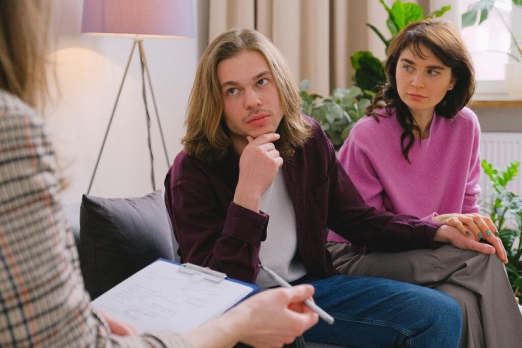 Fotografia de plano médio mostrando um homem de cabelos longos e camisa vinho com a mão no queixo e uma mulher de blusa rosa sentados em um sofá, olhando atentamente para uma terapeuta em primeiro plano que segura uma prancheta.