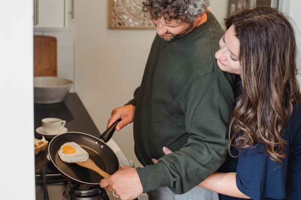 Fotografia de plano médio de uma mulher abraçando o parceiro por trás com carinho, enquanto ele prepara um ovo frito na frigideira sobre o fogão na cozinha.