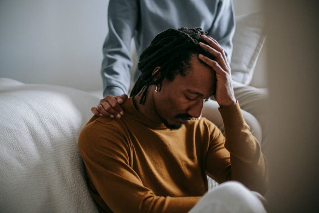 Fotografia de plano médio mostrando um homem negro com dreadlocks, sentado no chão e apoiando a cabeça na mão com uma expressão cansada, enquanto outra pessoa ao fundo, de moletom cinza, repousa a mão carinhosamente em seu ombro.