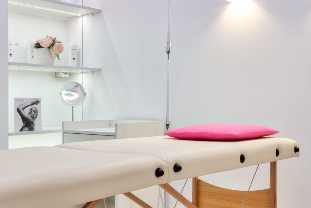 Fotografia em plano médio de uma maca de massagem dobrável estofada em cor bege claro, com uma almofada rosa sobre a cabeceira, posicionada em um ambiente de paredes brancas com prateleiras iluminadas por fitas de LED ao fundo, contendo cosméticos e um arranjo de flores.