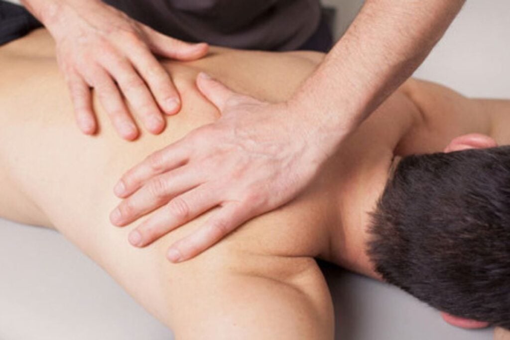 Homem recebendo massagem nas costas em maca, com profissional aplicando movimentos manuais na região dos ombros.